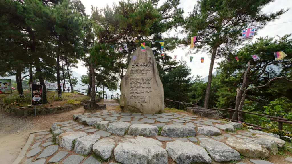 충청북도 괴산군 산막이옛길 입구의 기념비석과 숲길. 나무와 깃발 장식이 어우러진 자연친화적인 산책길 시작점 모습.